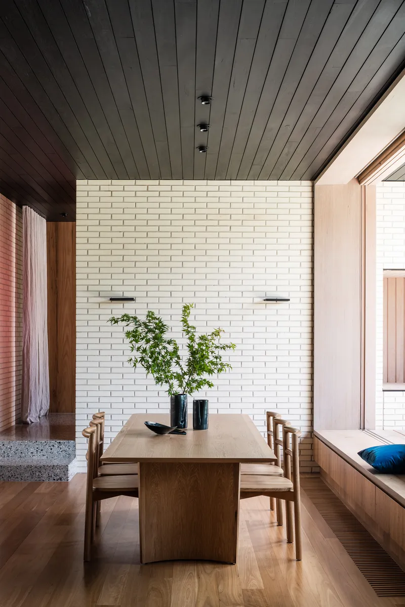 Dining space with timber table and chairs against cream brick wall under black timber ceiling with skylight