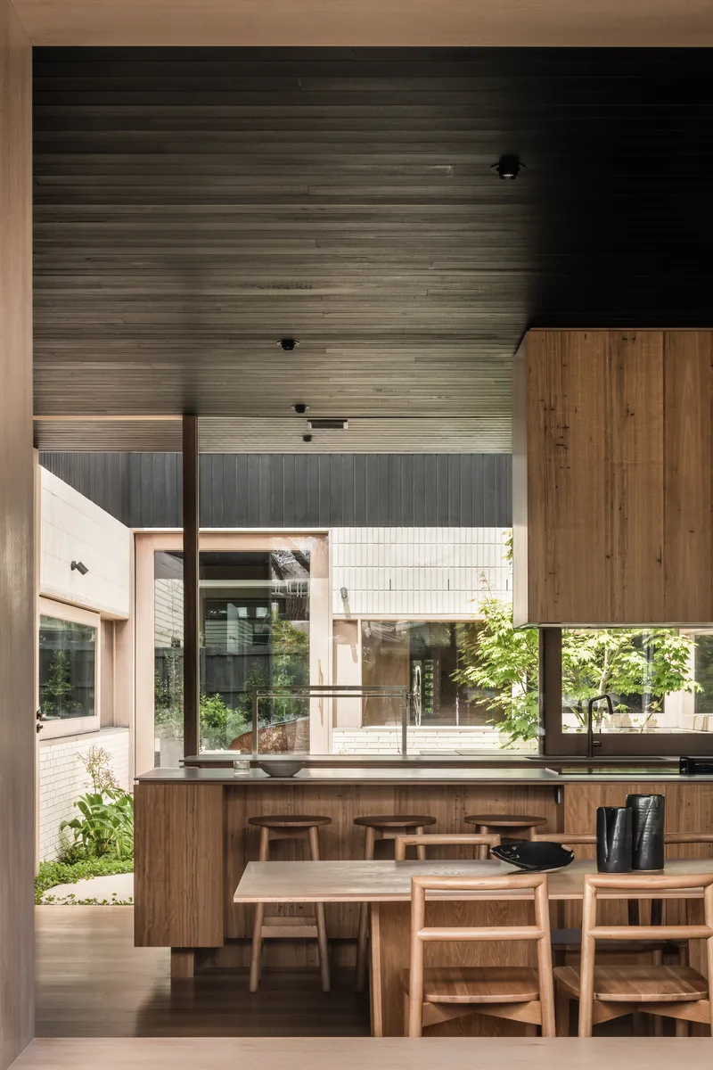 Kitchen and dining area with timber cabinetry and stools beneath high timber ceiling looking onto planted courtyard
