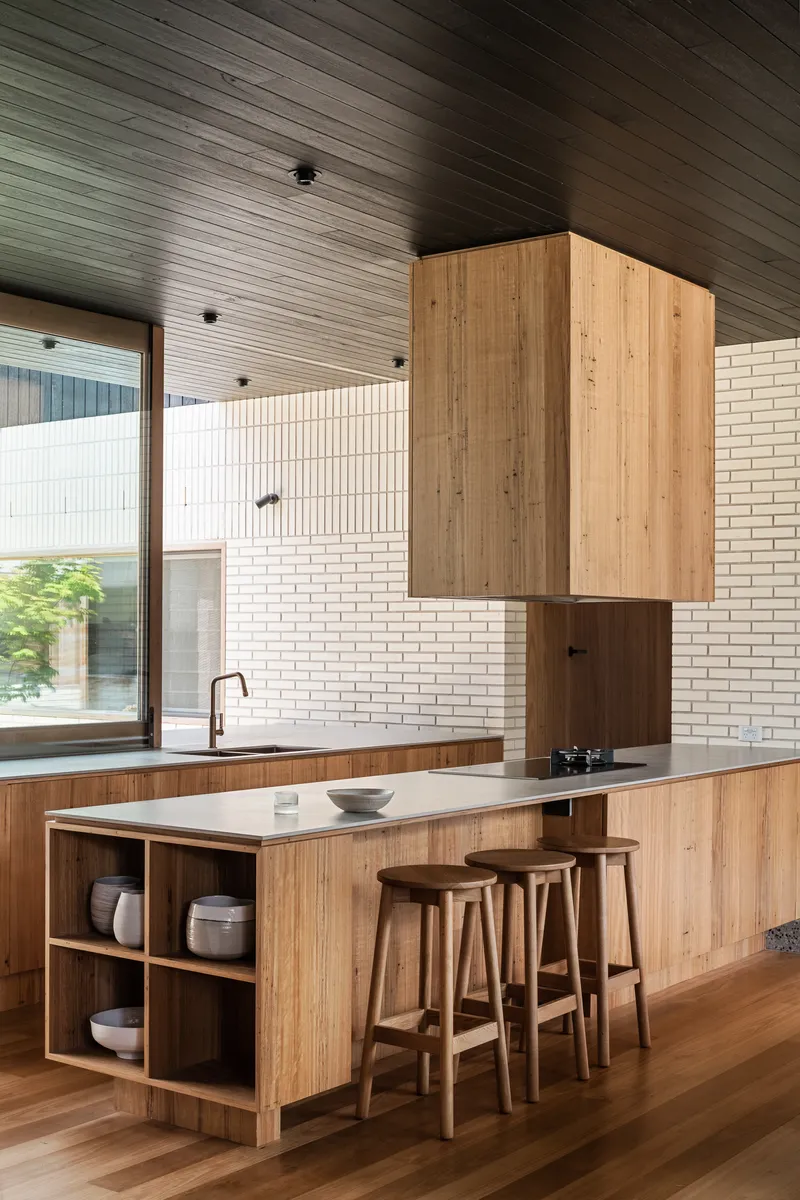 Kitchen island with timber cabinetry and three stools beneath a suspended timber hood and slatted ceiling