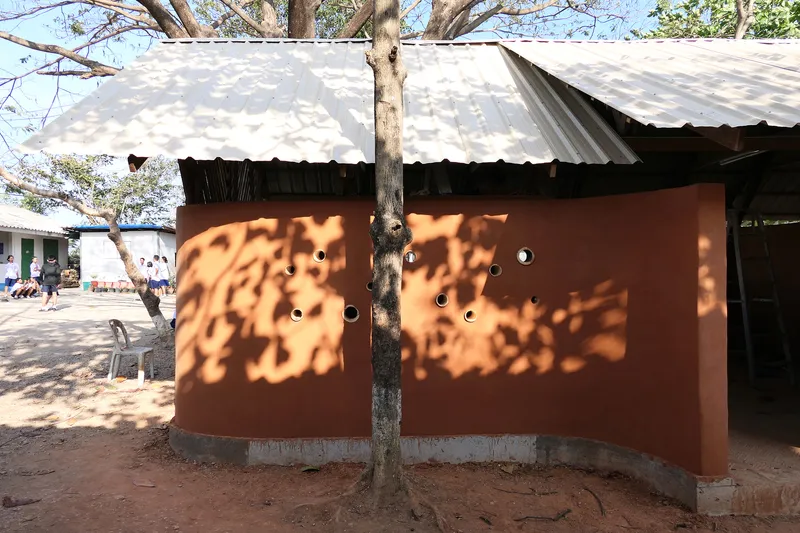 Circular perforations in rammed earth wall with dappled tree shadow and corrugated metal roof overhang
