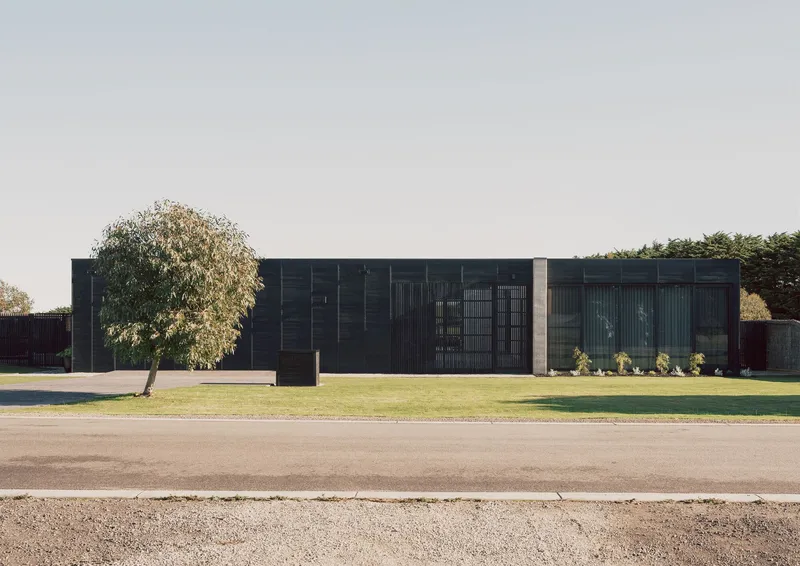 Street view of the dark timber facade with vertical louvres beside a mature tree on the lawn