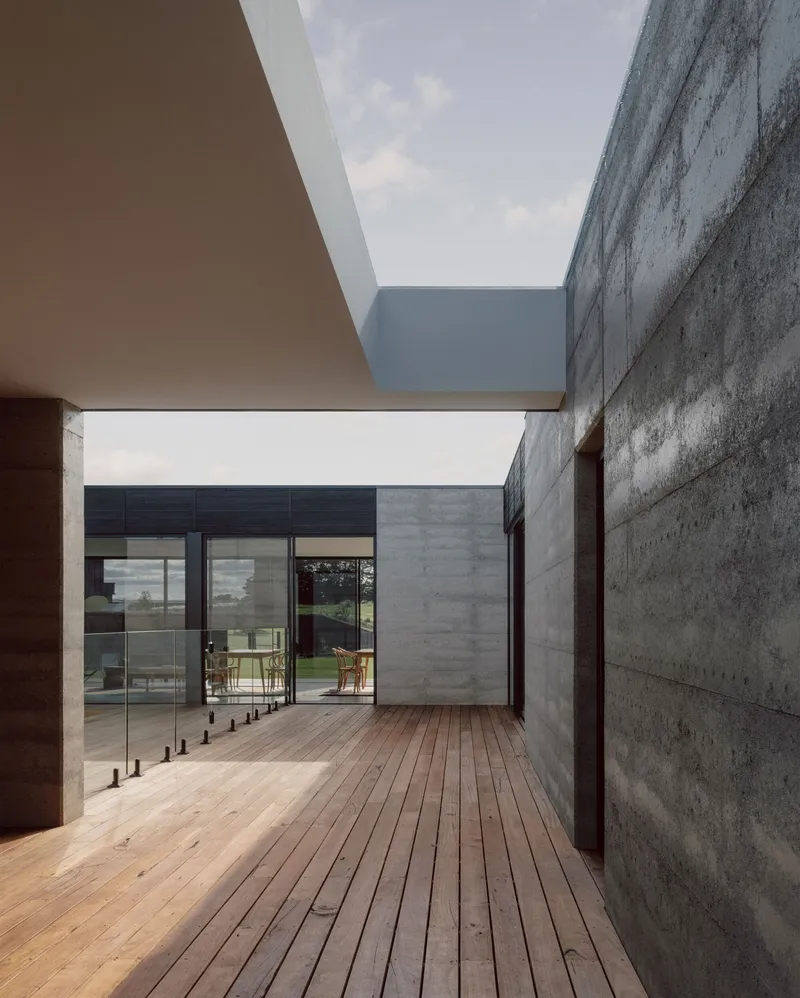 Timber deck courtyard framed by boardformed concrete walls and a cantilevered overhang under blue sky