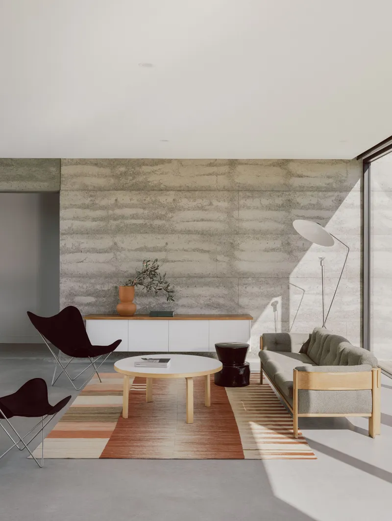 Interior view showing butterfly chairs and low table on striped rug beside boardformed concrete wall