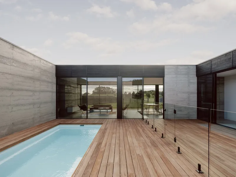 Courtyard pool with timber deck and glass balustrade framed by boardformed concrete walls under cloudy sky