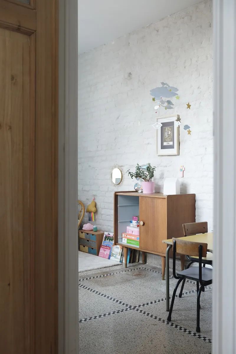 View through timber doorway into a playroom with whitewashed brick wall and mid-century cabinet