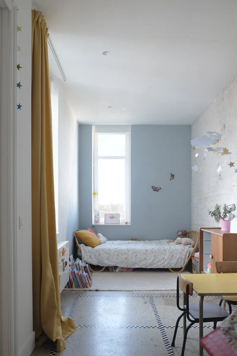 Narrow bedroom with blue accent wall and terrazzo flooring divided by yellow curtain