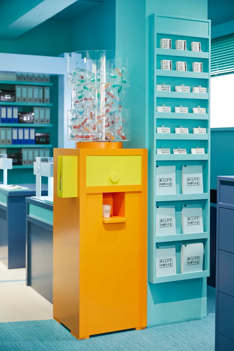 Yellow pedestal dispenser with transparent acrylic top filled with colorful objects beside turquoise shelving unit