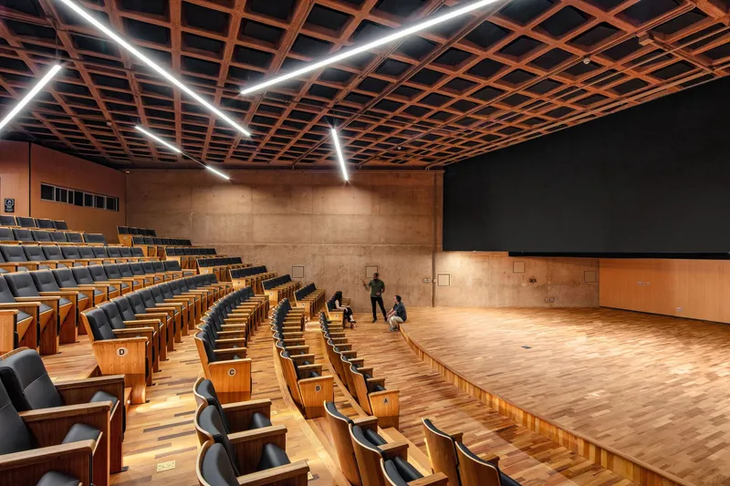 Auditorium with timber grid ceiling, linear lighting, tiered seating, and a wooden stage floor