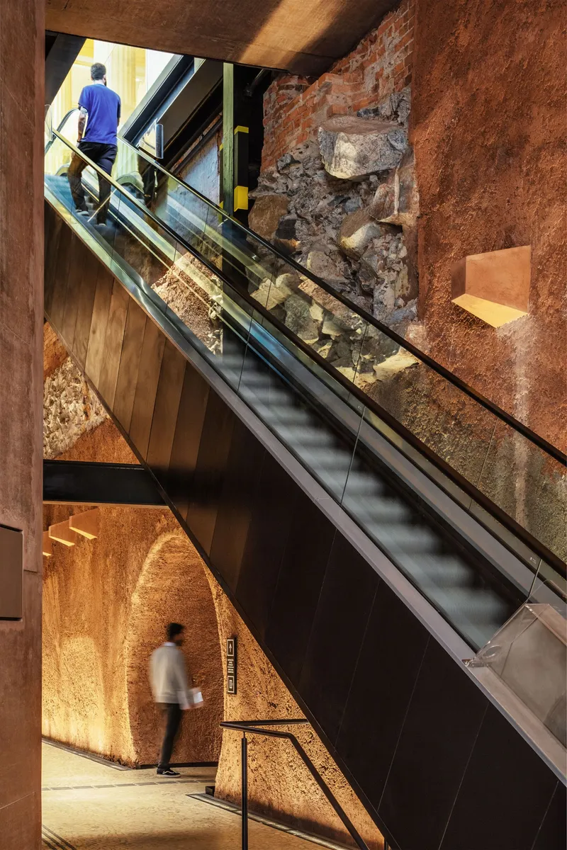 Escalator cutting through excavated earth and exposed brick walls with visitors ascending through layered space