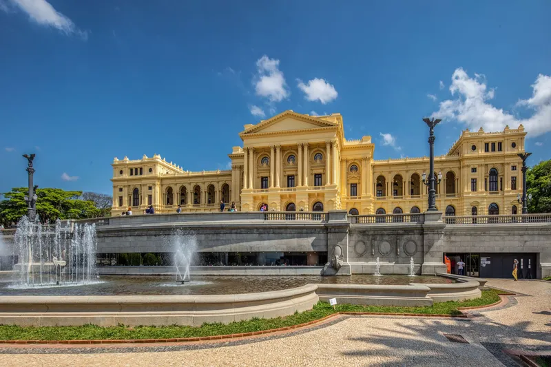 Circular fountain plaza with arched bridge leading to a columned entrance under blue sky