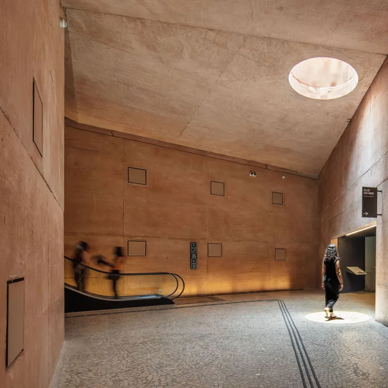 Rammed earth lobby with circular skylight, escalator, and blurred figures in motion across terrazzo floor