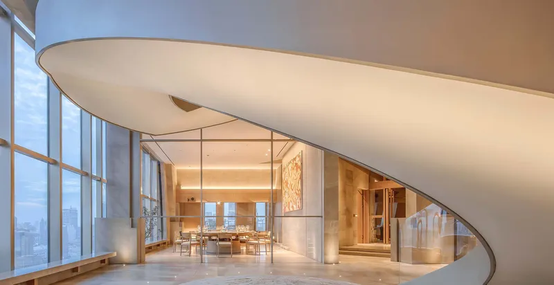 Curved white staircase with underside view showing smooth soffit beside tall curtain wall glazing