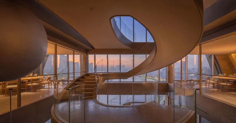 Helical staircase rising through two dining levels with floor-to-ceiling windows overlooking the cityscape at dusk