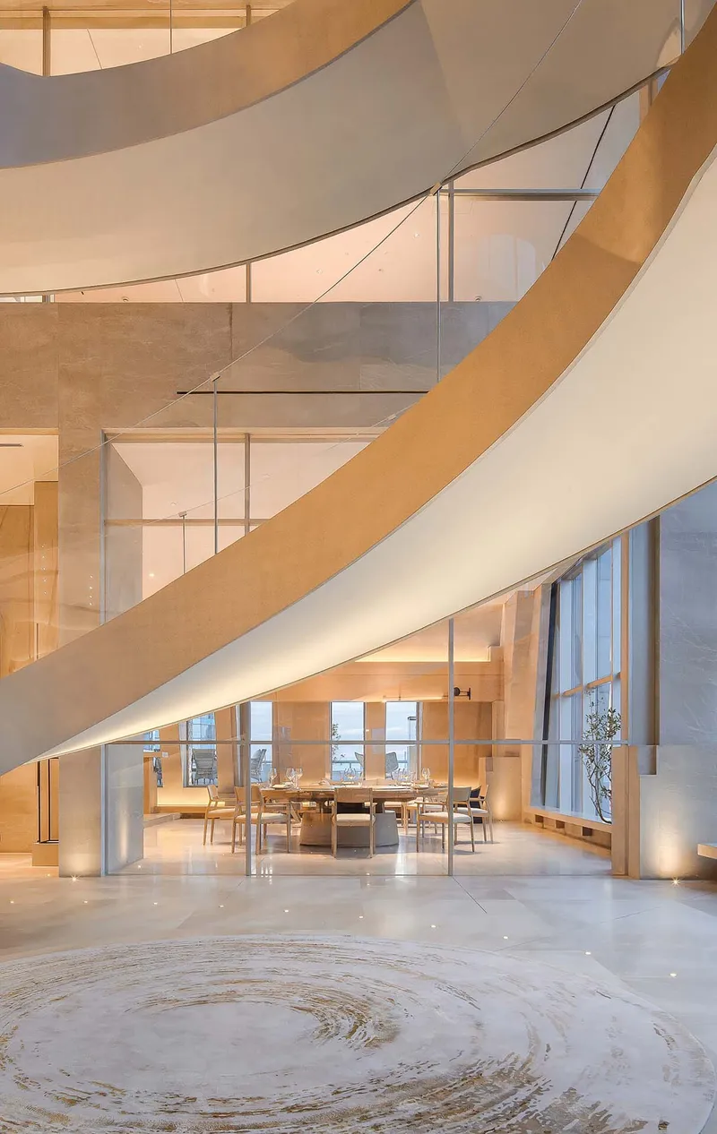 View beneath sweeping wood-clad stairs into a dining area with pale stone flooring and pendant lights