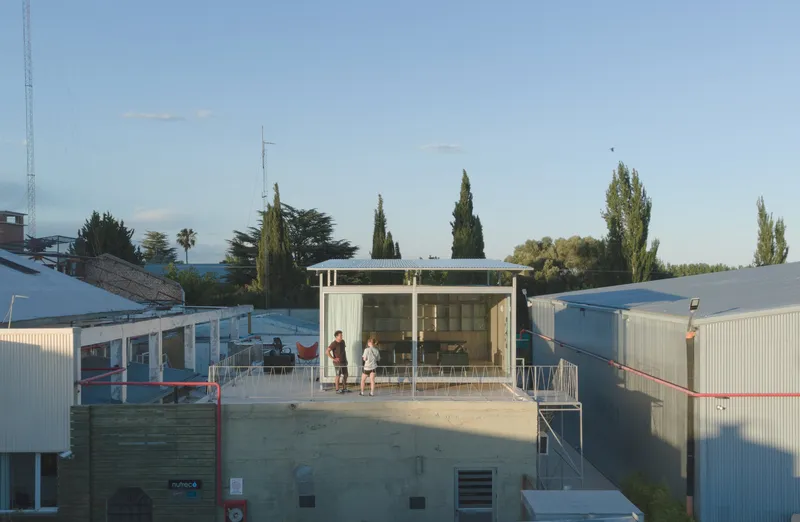 Elevated glass-walled pavilion on a concrete base surrounded by corrugated metal structures and cypress trees