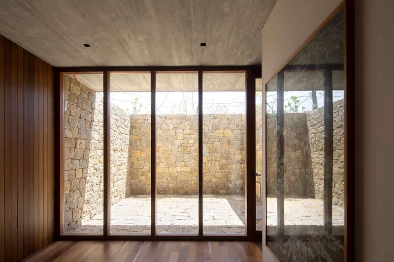 Interior view through floor-to-ceiling glazing framing the stone-walled courtyard in afternoon light