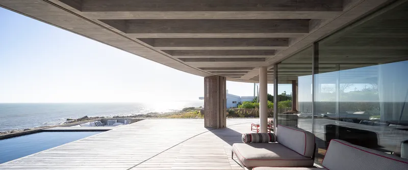 Covered terrace with concrete ceiling beams and perforated timber screens overlooking an infinity pool and coastline