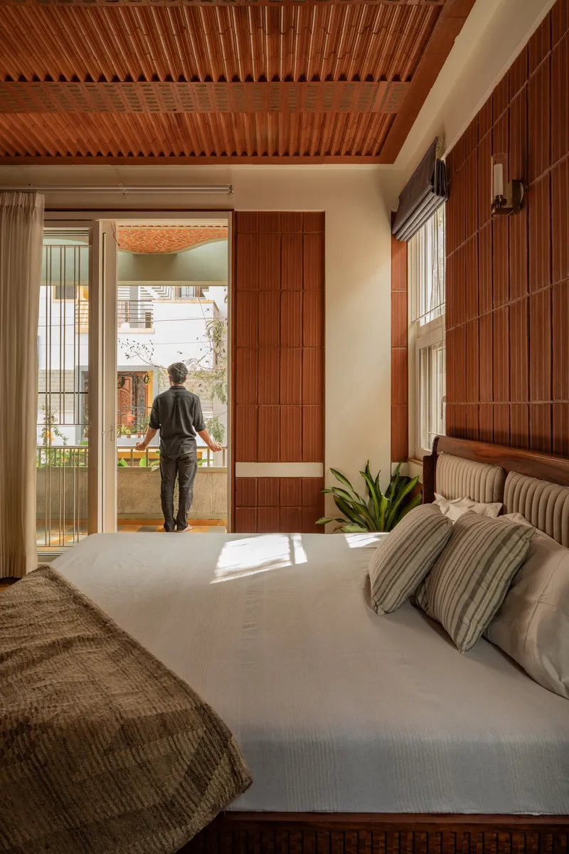 Bedroom with timber-slatted ceiling and wall as a figure opens glazed doors to a sunlit terrace