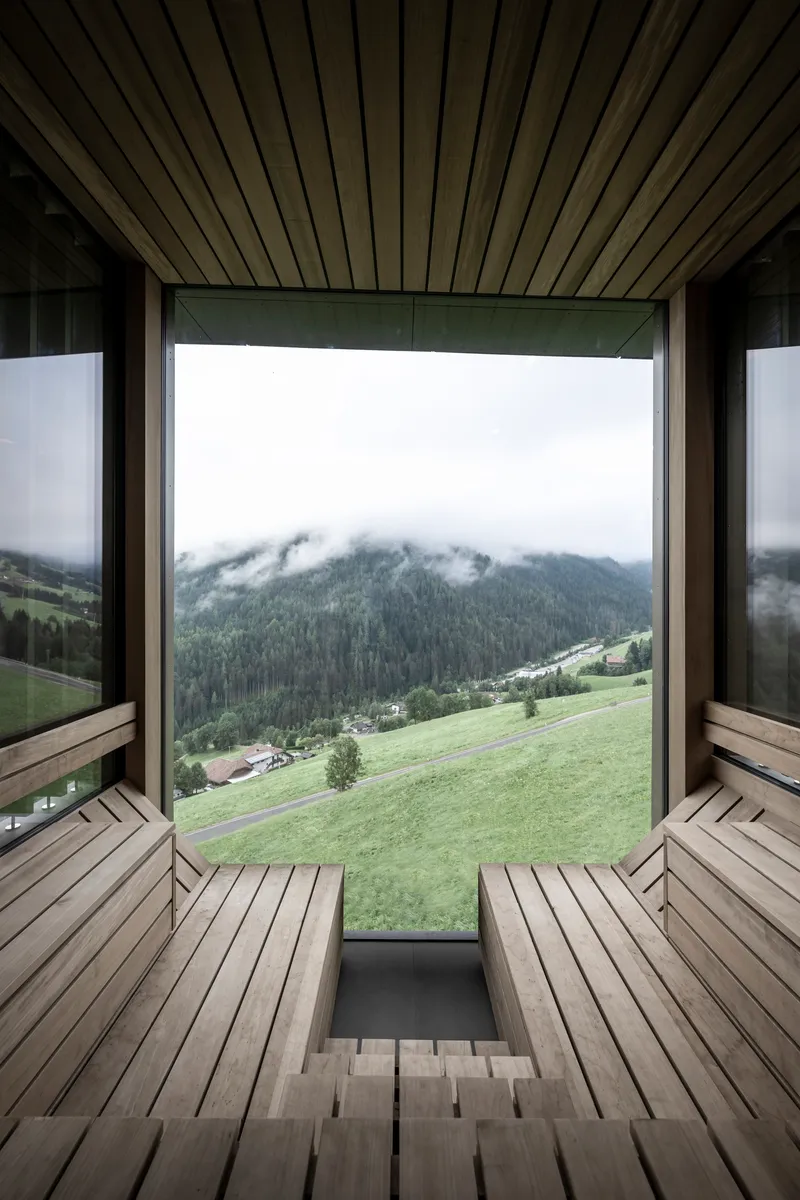 Timber sauna interior with slatted benches facing a picture window overlooking green meadows and mist