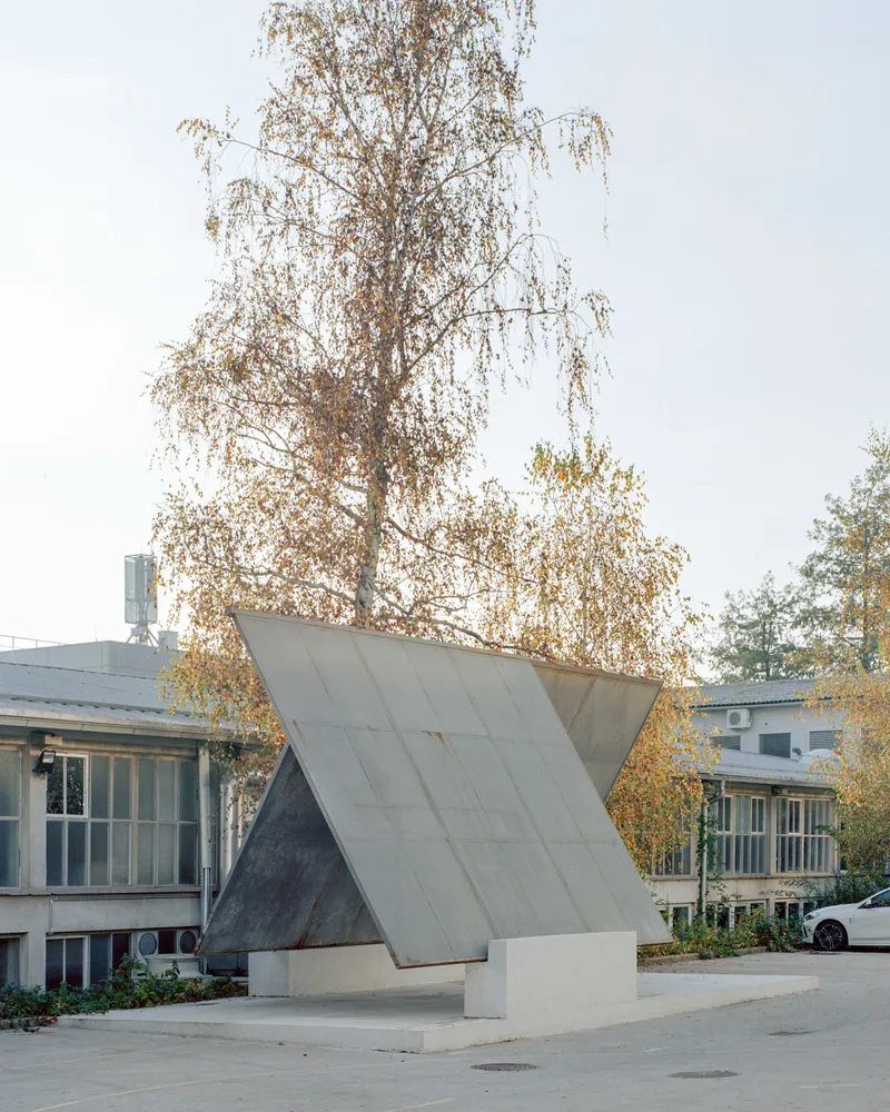Concrete sculptural element with tilted planes standing in the paved courtyard beside a birch tree