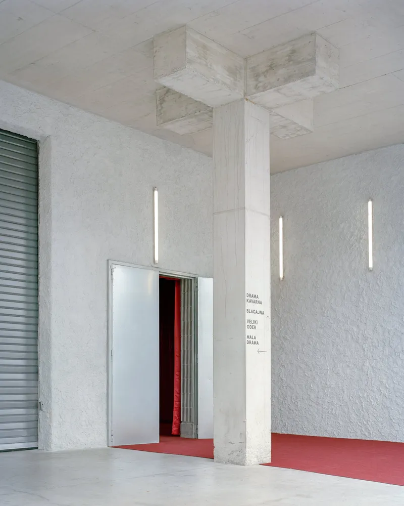 Entry column with textured white plaster walls, vertical tube lighting and red carpet leading through metal doors