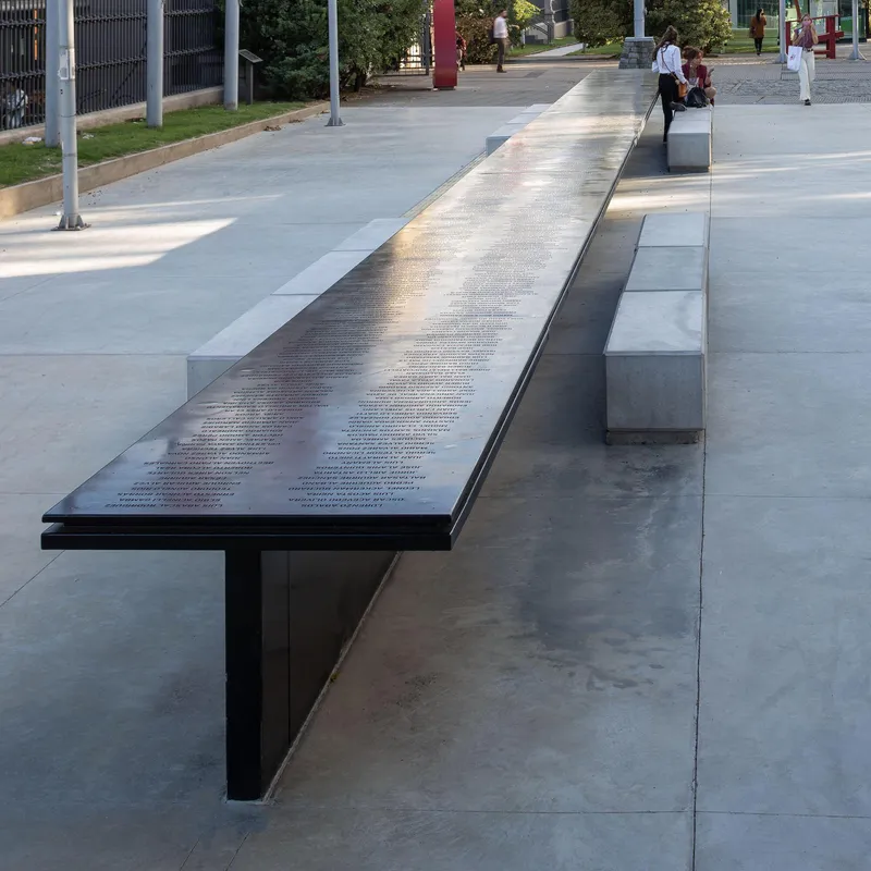 Long black metal table with etched text flanked by concrete benches in a public plaza