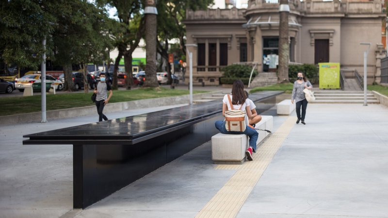 Black metal memorial table with engraved text beside concrete seating blocks and pedestrians