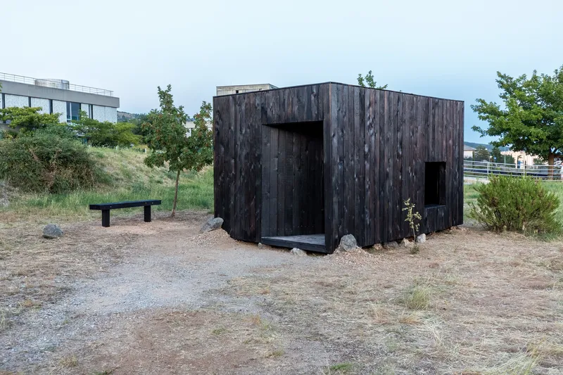 Small blackened wood pavilion with two openings on cleared ground next to a wooden bench
