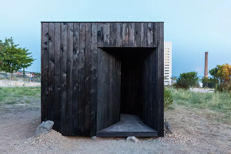 Head-on view of the charred vertical timber facade with trapezoidal entry and gravel approach