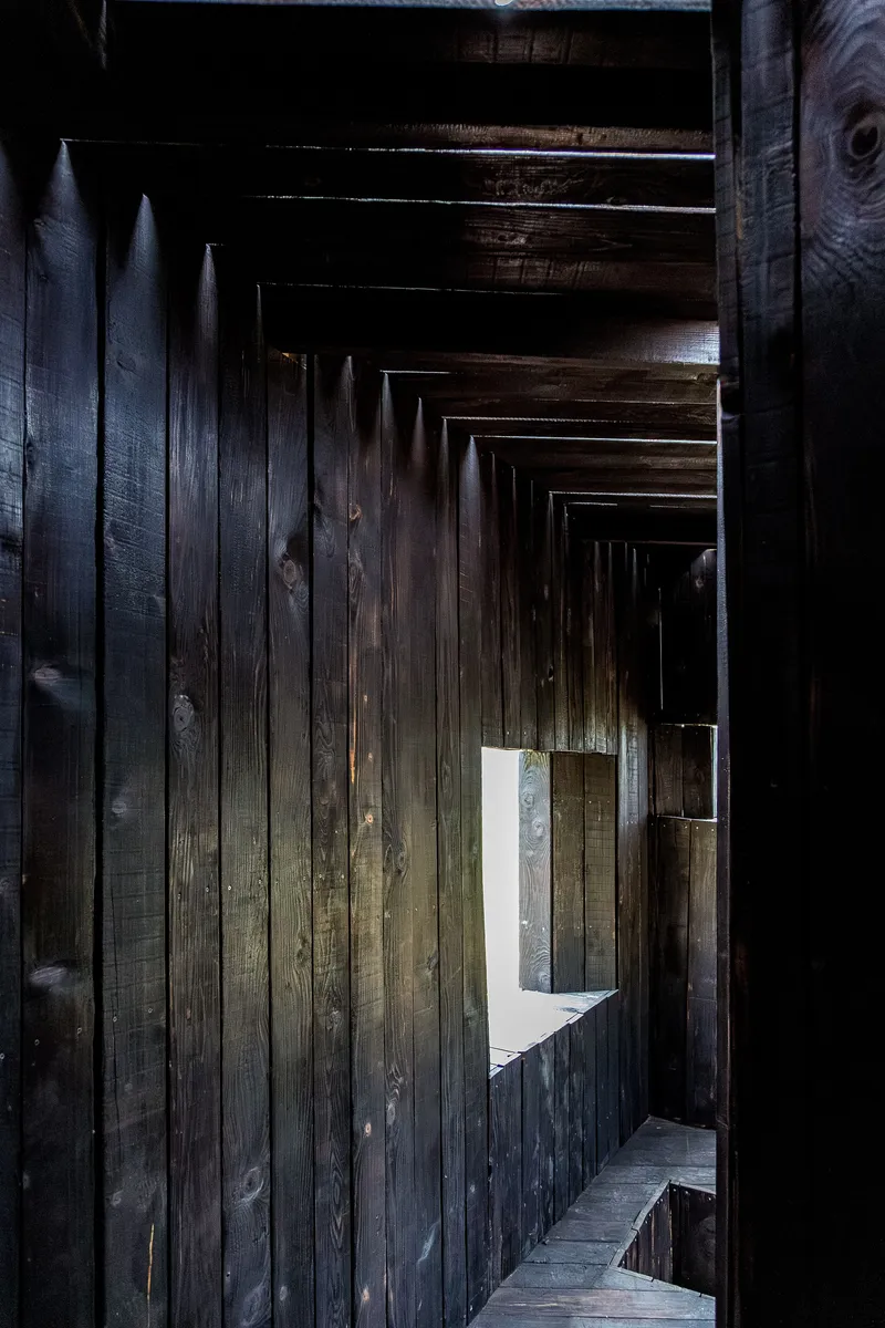 Interior passage with charred wood plank walls and exposed joists leading toward a window opening