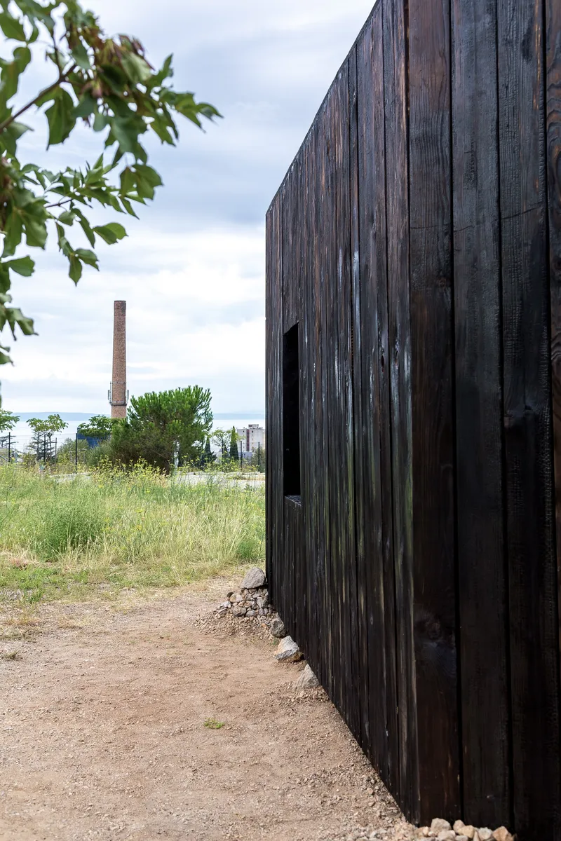 Charred vertical timber cladding with a window slot facing a gravel path and industrial chimney