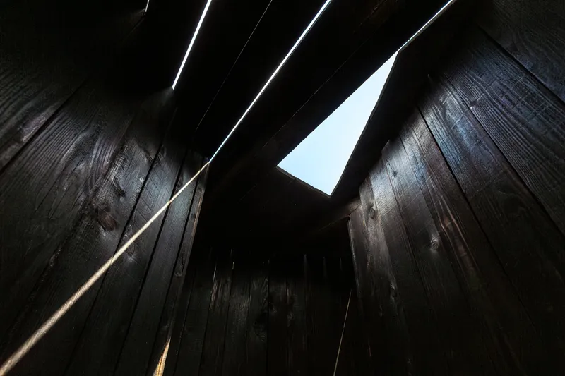 Upward view through a triangular skylight opening between charred timber walls with light streaks