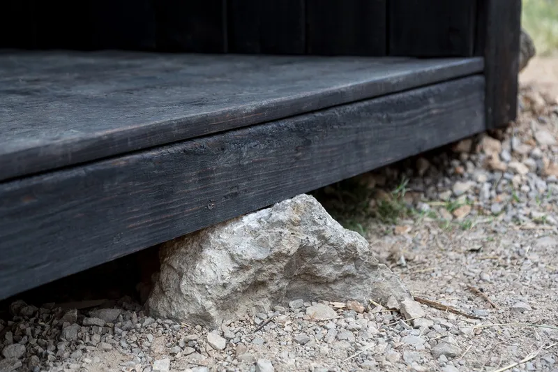 Detail of charred timber threshold meeting gravel ground with a single weathered stone beneath