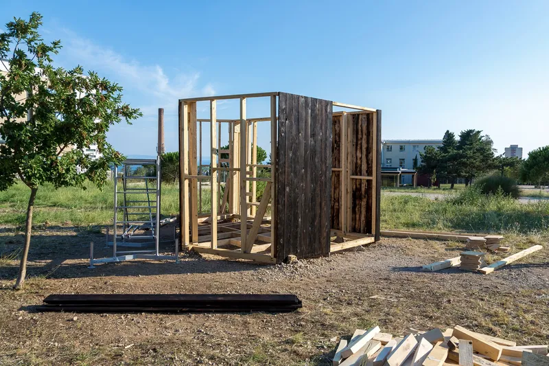 Timber frame structure under construction on a gravel site with exposed studs and weathered cladding panels