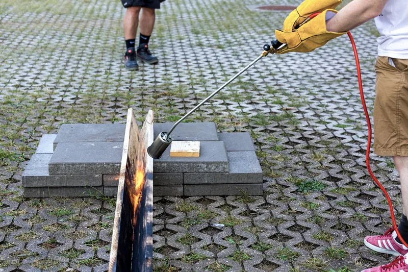 Hands using a torch to char timber samples arranged on concrete pavers in a plaza