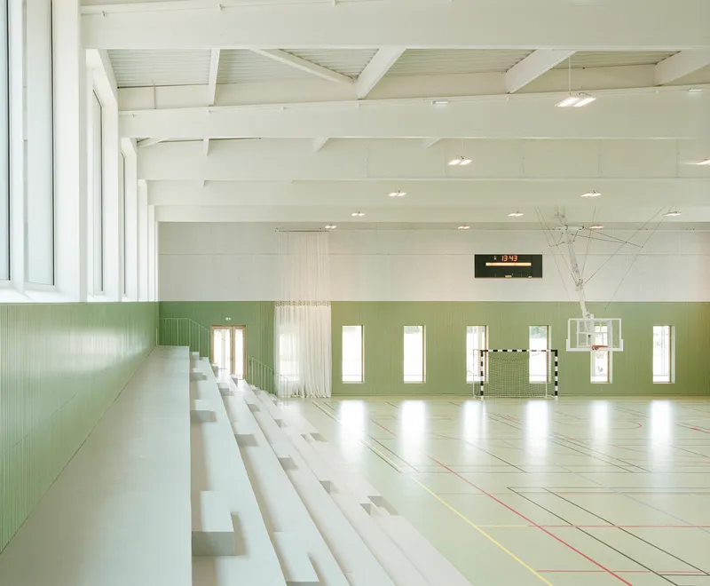 Indoor sports hall with white bleacher seating, green tiled wall and exposed beam ceiling structure