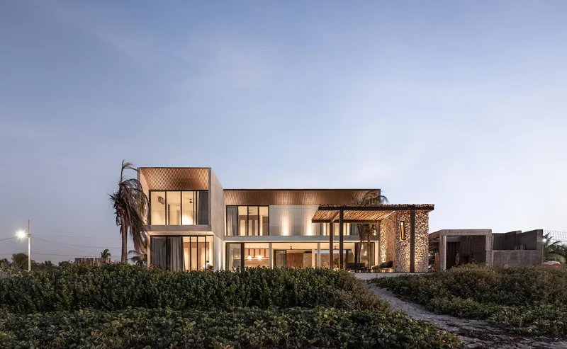 Street view of the residence with timber-clad volumes and coastal vegetation at twilight