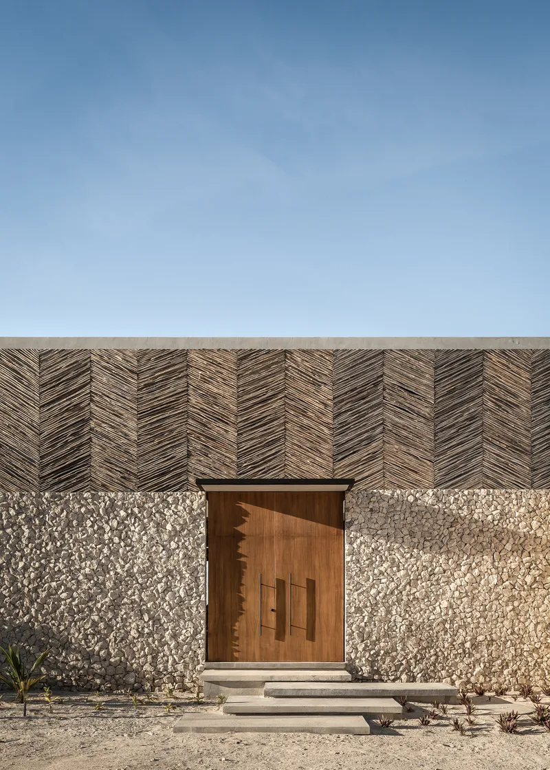 Entrance facade with herringbone timber cladding above stone aggregate walls and timber pivot door under clear sky