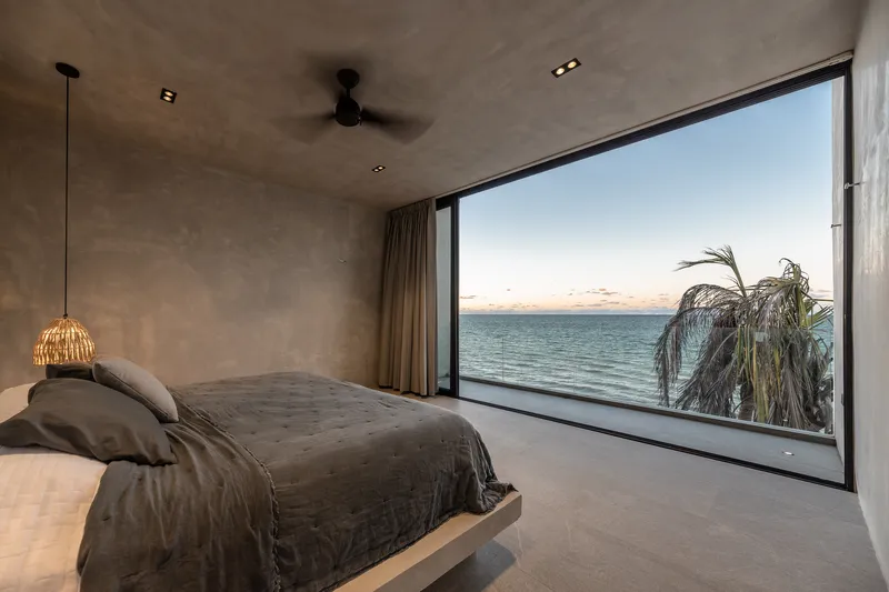 Bedroom with full-height glazing framing ocean view and palm fronds at dusk