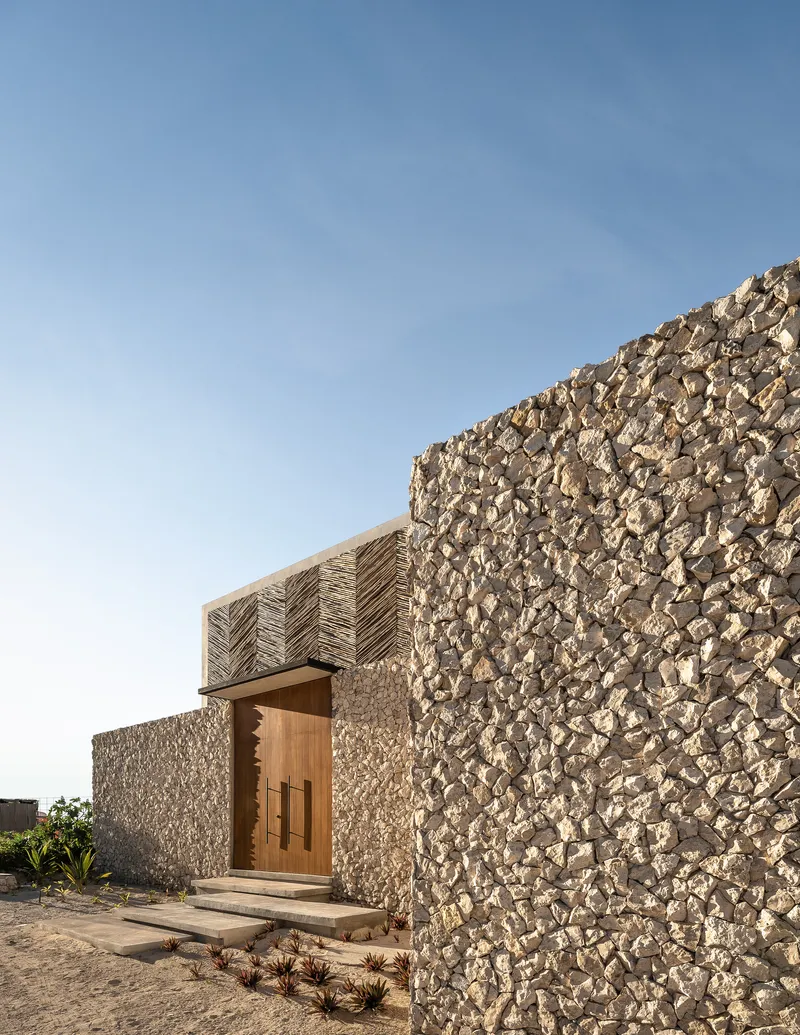 Corner view of stone aggregate walls meeting herringbone timber roof with timber entry door and low plantings