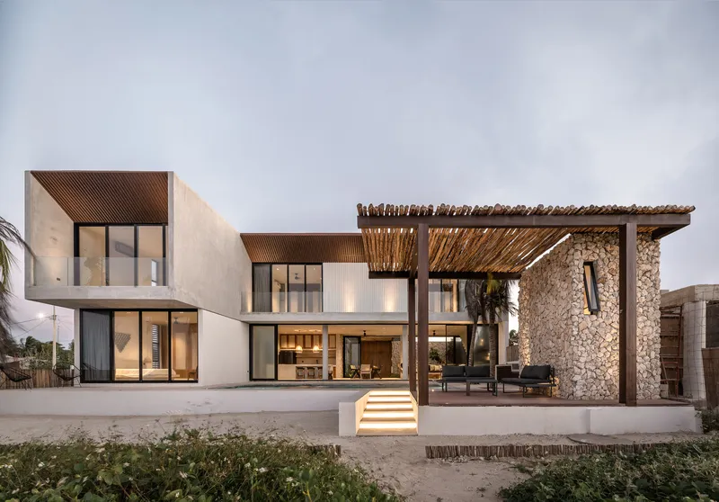 Rear facade showing white rendered volumes with a gabion wall pergola and illuminated entry steps at dusk