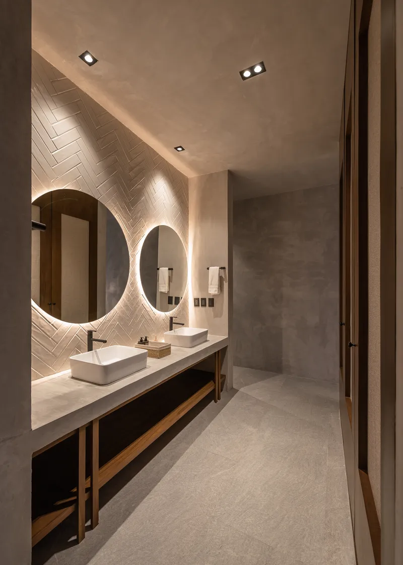 Bathroom vanity with backlit circular mirrors and herringbone tile wall under recessed lighting