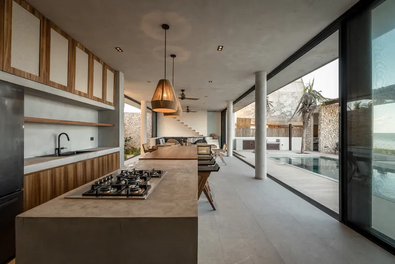 Open kitchen with concrete island and floor-to-ceiling glazing overlooking a pool and gabion walls
