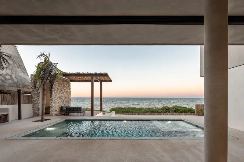 Pool terrace framed by concrete beams with ocean view and thatched pavilion at dusk