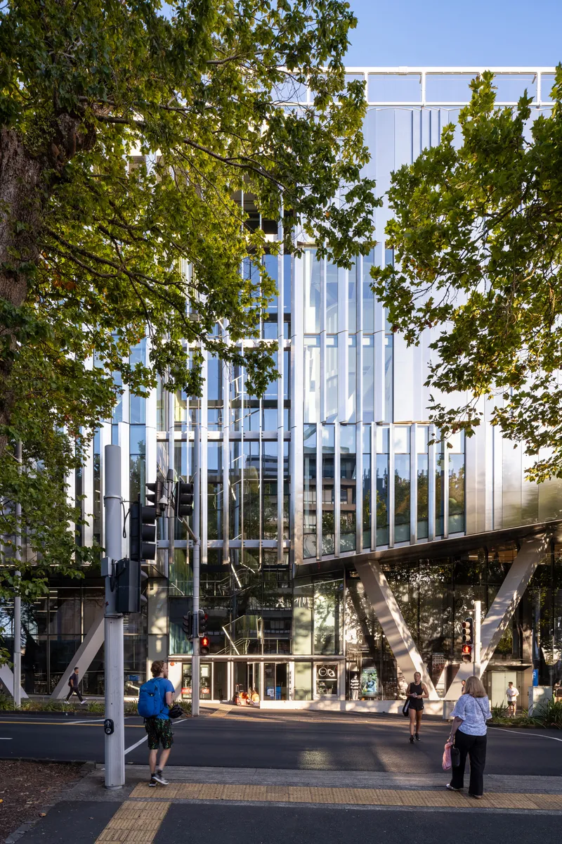 Vertical white louvred facade framed by mature tree canopy and angled structural columns at ground level
