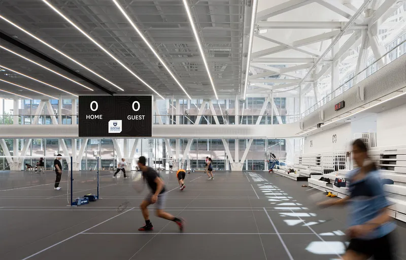 Multi-court sports hall with white diagonal bracing and digital scoreboard during active play