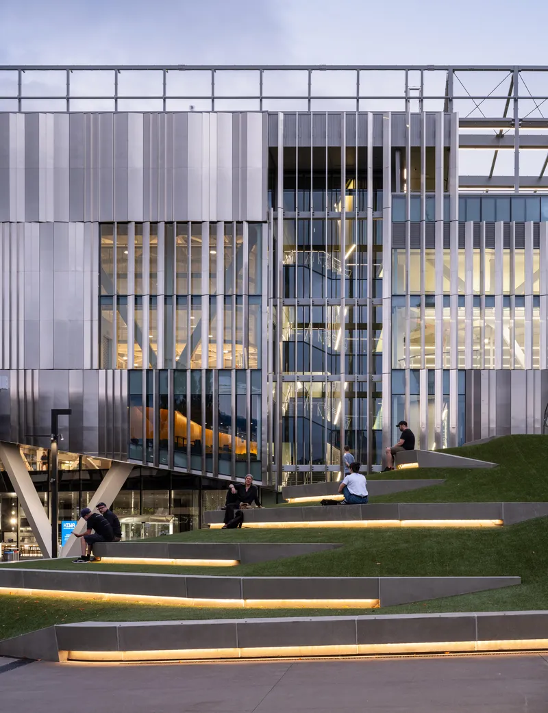 Exterior view of translucent facade with vertical fins and stepped lawn with visitors at dusk