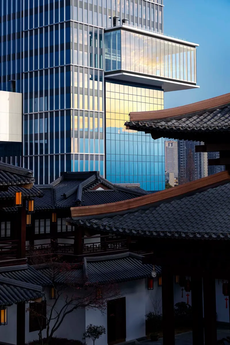 Glass tower with cantilevered volume rising behind traditional timber and tile roofed buildings at dusk