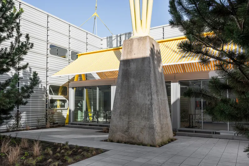 Exterior courtyard with tapered concrete monument, yellow and white striped fabric canopy, and corrugated metal cladding