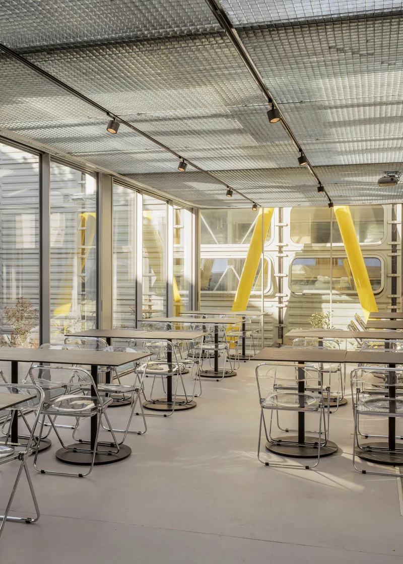 Dining space with mesh metal ceiling, transparent chairs, and yellow diagonal bracing seen through glass partition
